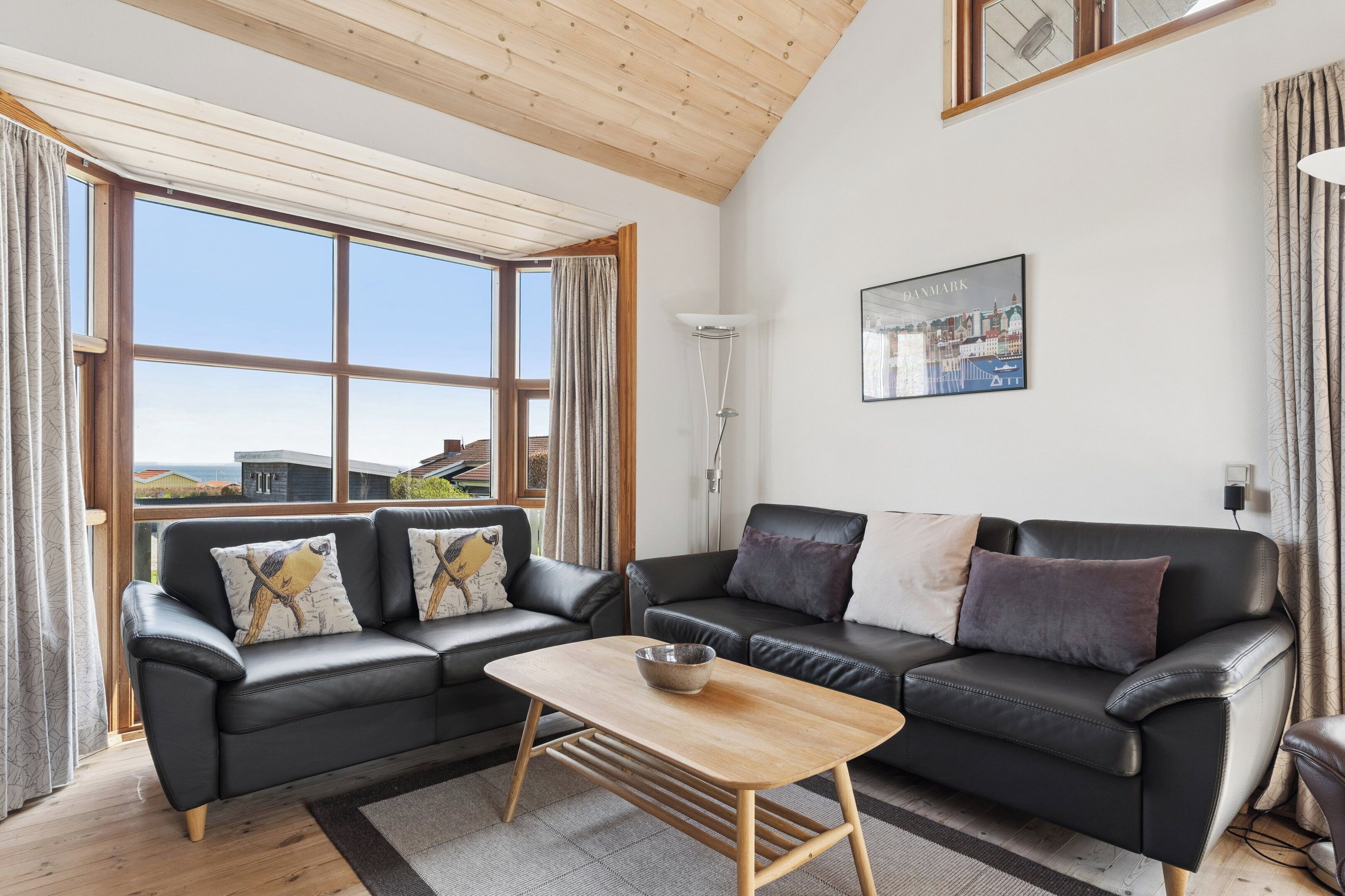 Een moderne woonkamer met twee zwarte leren banken, een houten salontafel en grote ramen met uitzicht op gebouwen.