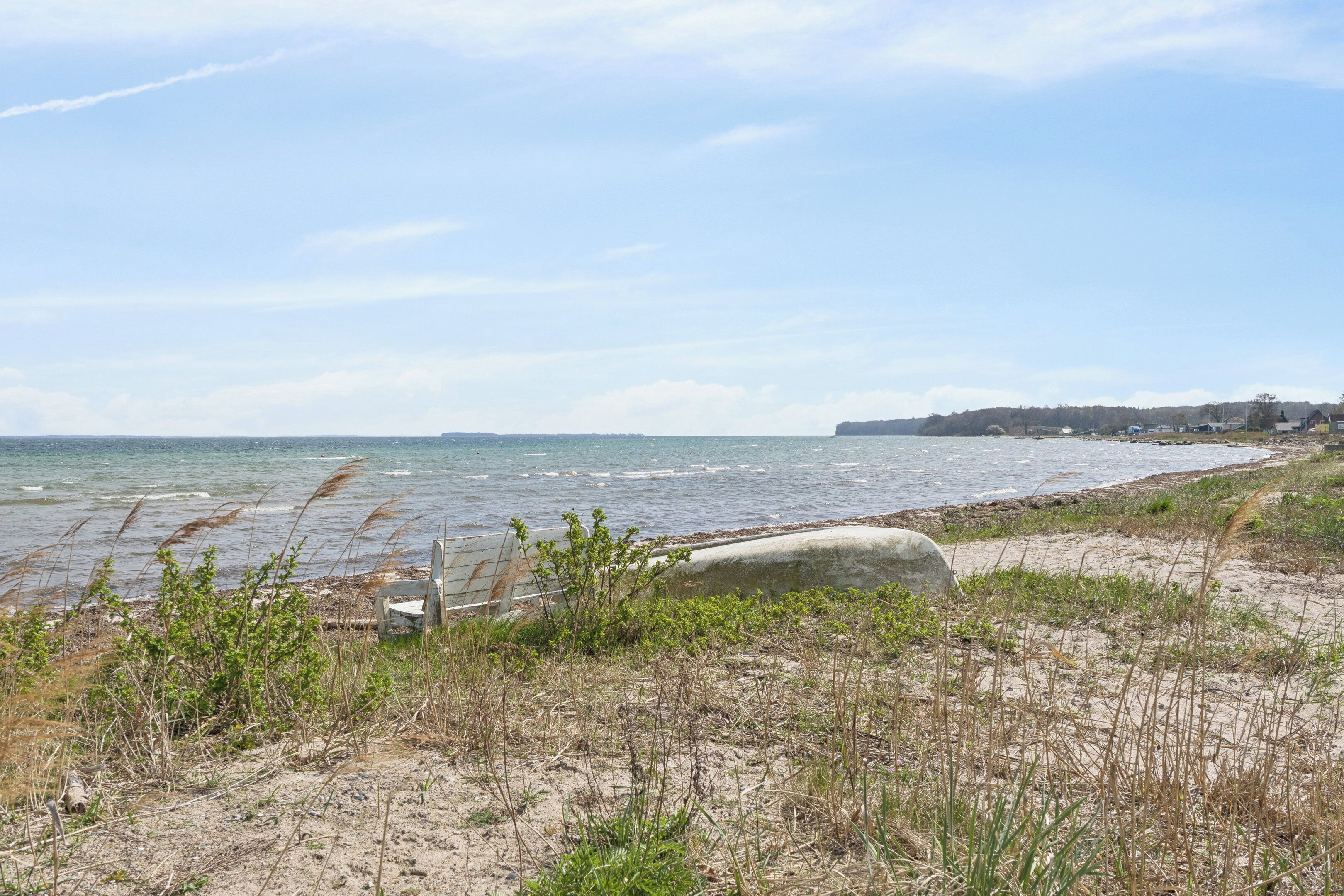 Ett kustlandskap med en sandstrand, en betongbarriär och en avlägsen kustlinje med byggnader.