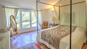 A four-poster bed with a patterned bedspread, a standing mirror, a bedside table with a lamp, and a view of the outdoors through a glass door. - La Garrigue de Gordes (Gordes)