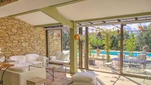 A spacious living area with a stone wall, white furniture, a glass coffee table, and a view of a pool and garden. - La Garrigue de Gordes (Gordes)