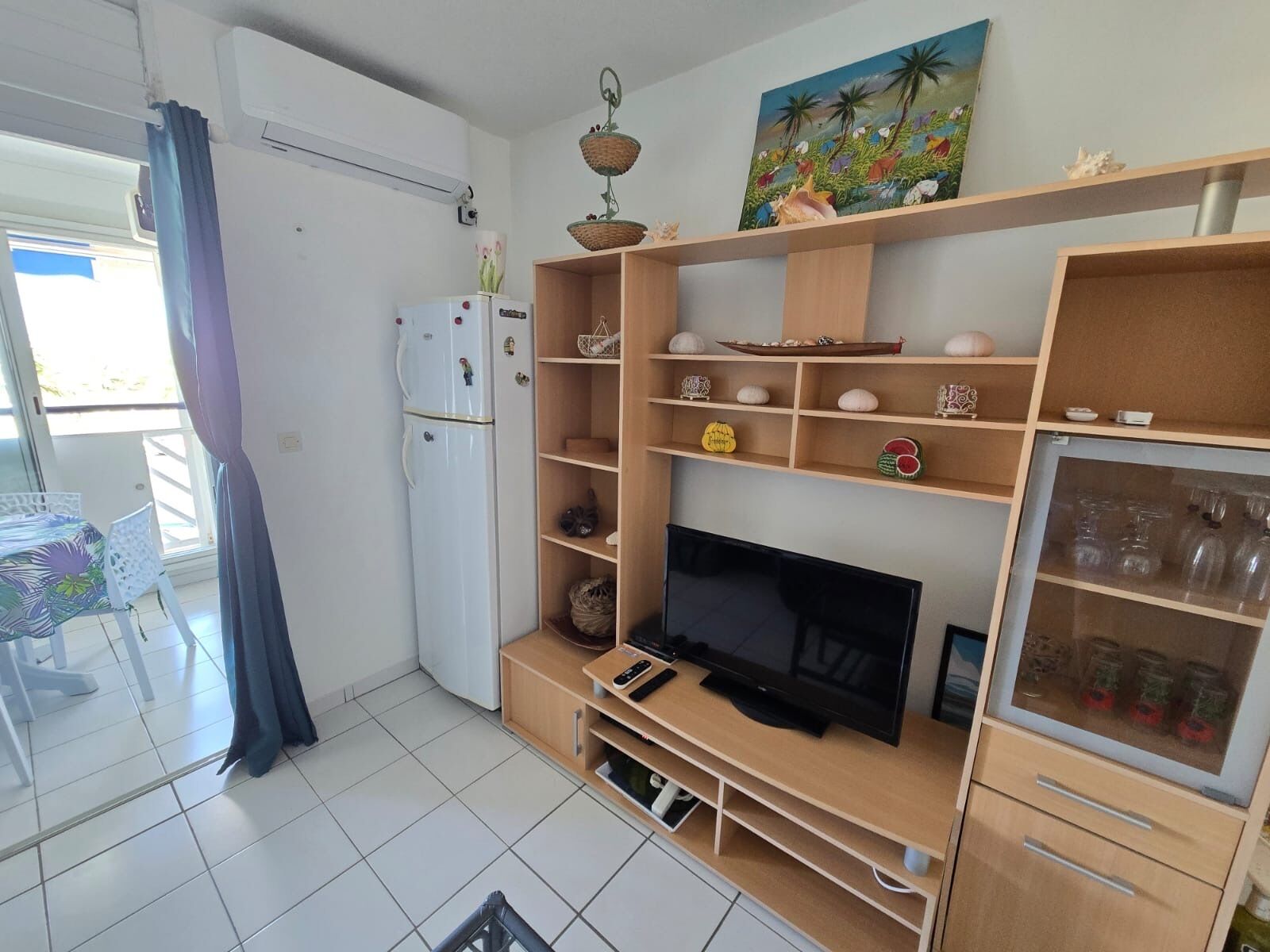 Una sala de estar con televisor, estanterías con adornos, refrigerador y un comedor con mesa y sillas.