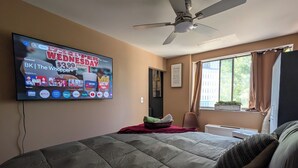 Een slaapkamer met een bed, een plafondventilator, een televisie aan de wand die een sportzender toont, en een raam met gordijnen.