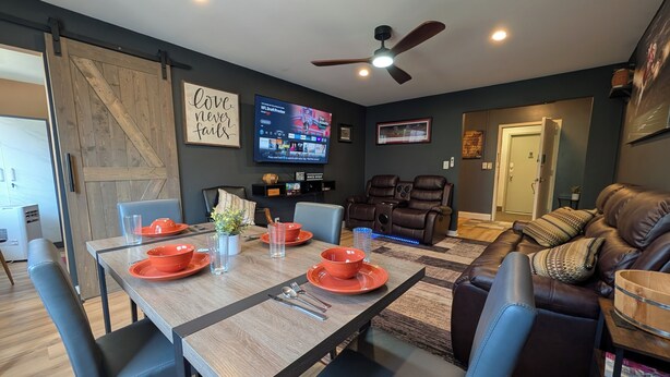 Een eethoek met een houten tafel, blauwe stoelen en oranje borden. Aan de muur hangt een televisie waarop sportbeelden te zien zijn.