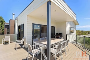 Une maison moderne dotée d’un patio couvert, d’un coin repas avec des meubles blancs et d’un barbecue.