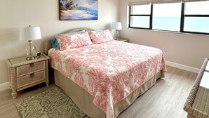 Una camera da letto con un letto, due comodini, una lampada da terra, un quadro, una finestra e vista sull'oceano.