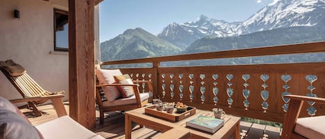 A wooden deck with a view of snow-capped mountains, a cushioned bench, and a table with a book and a tray of bread.