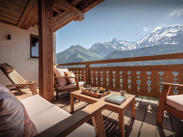 A wooden deck with a view of snow-capped mountains, a cushioned bench, and a table with a book and a tray of bread.