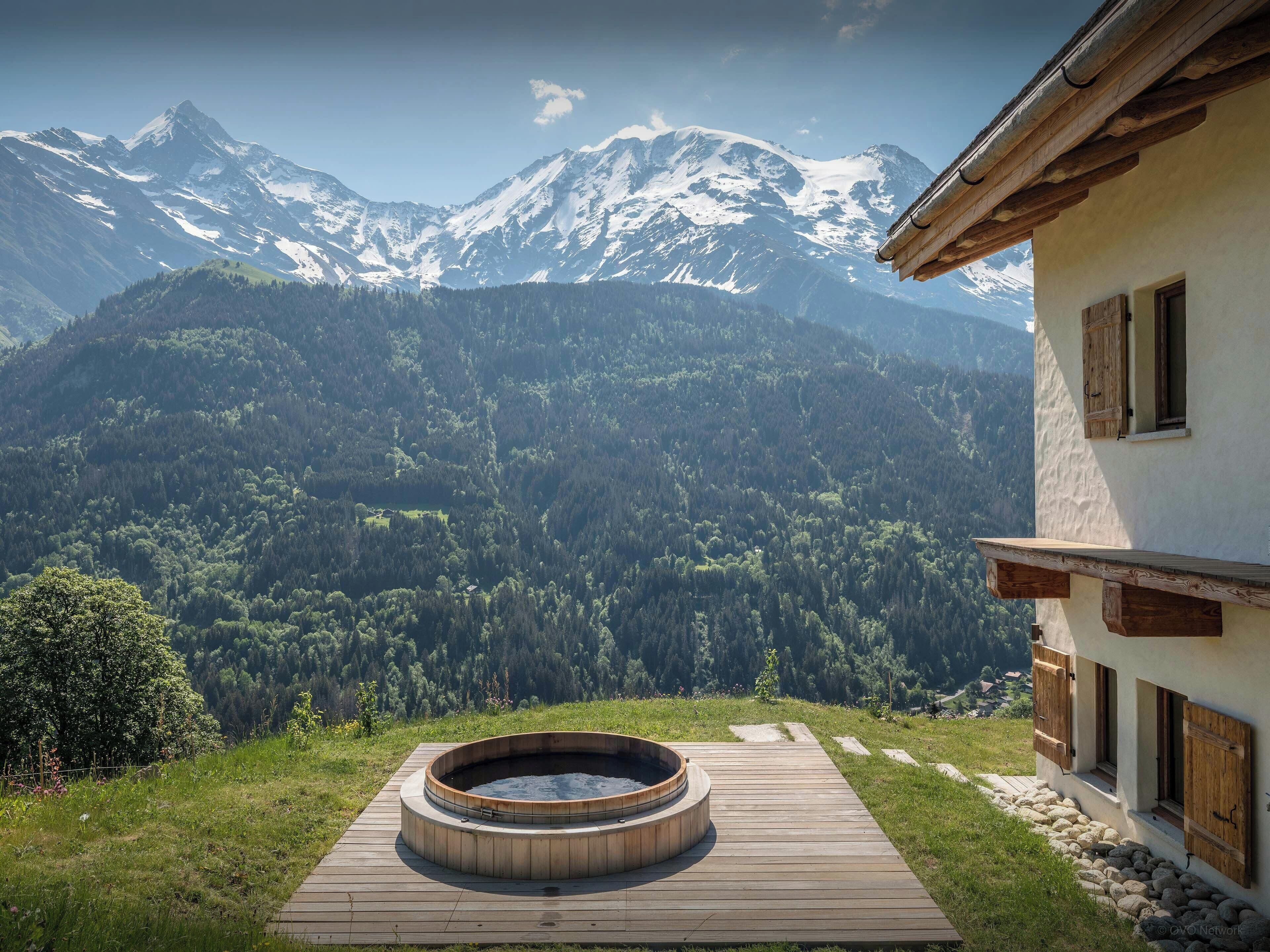 A wooden deck with a hot tub, a mountain view, and a building with wooden shutters.