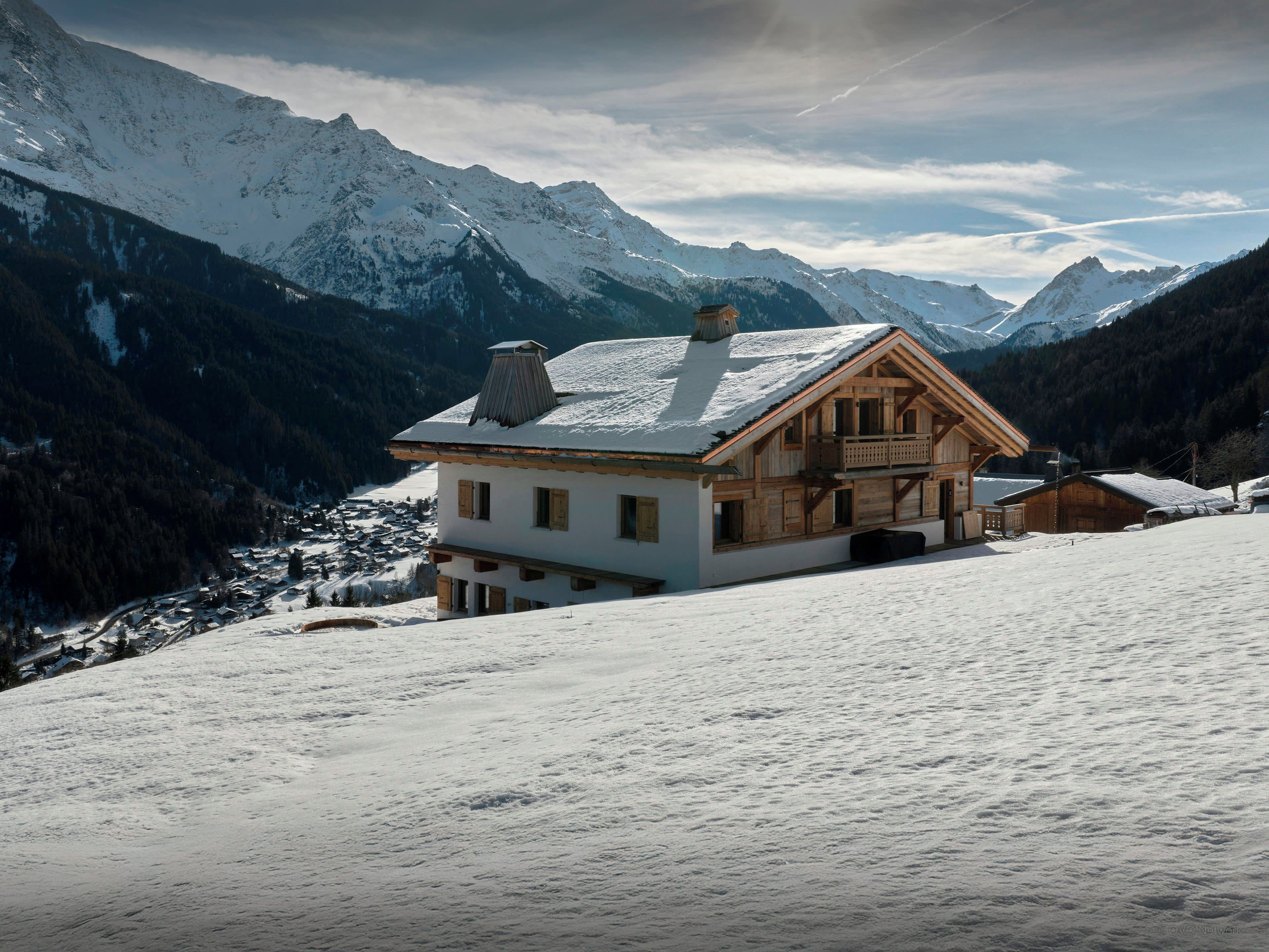Uma casa em estilo chalé com varanda, rodeada por montanhas cobertas de neve e um vale arborizado.