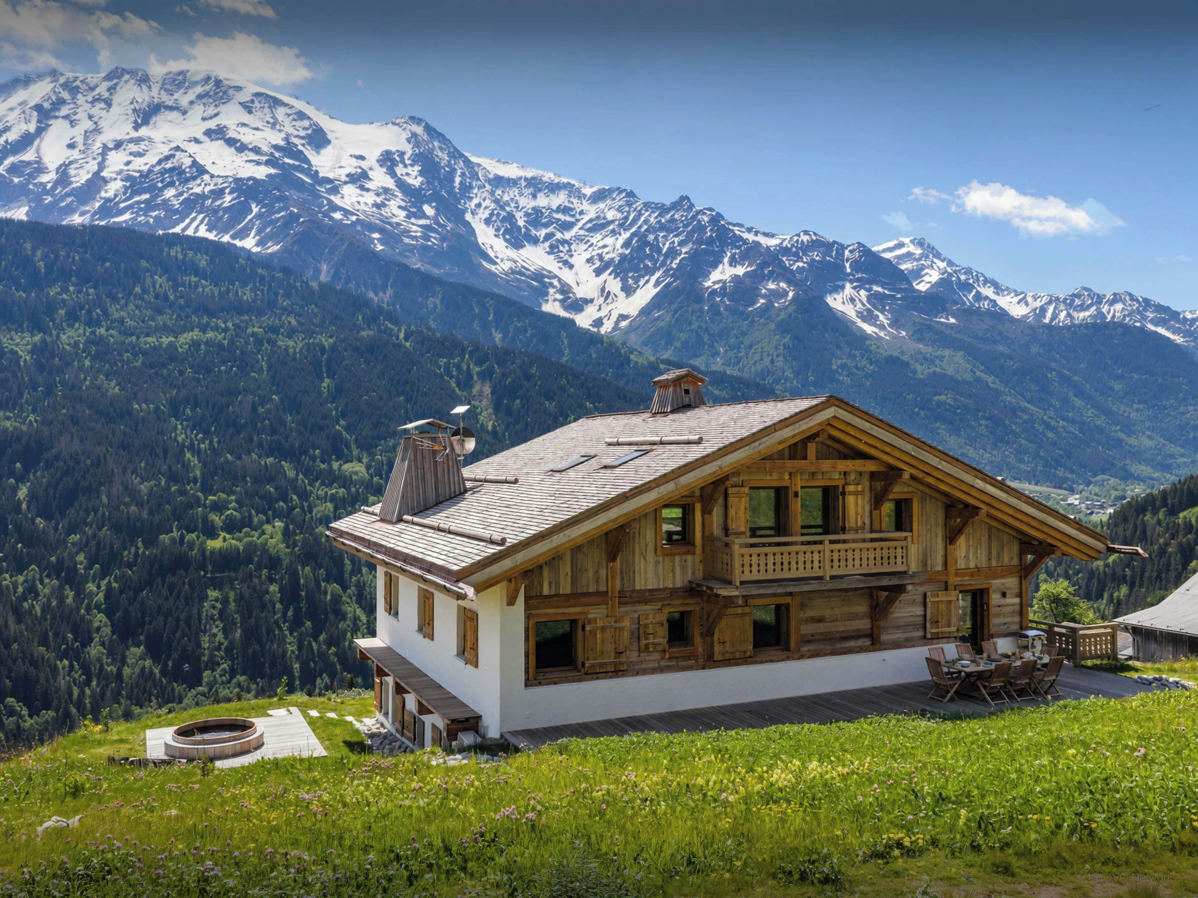 Un chalet de madera con balcón, con montañas nevadas y un valle boscoso al fondo.