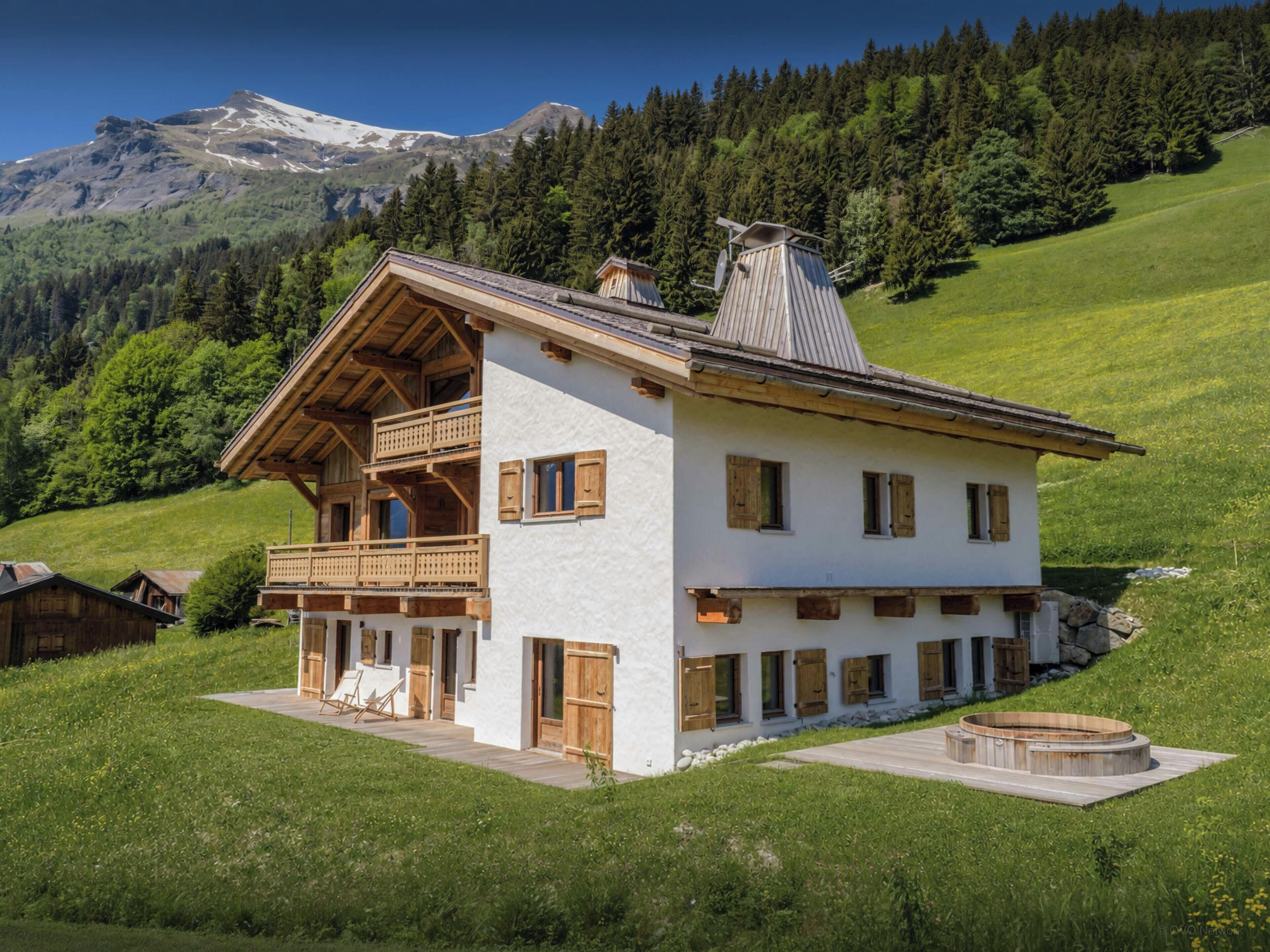 A white chalet with wooden balconies, a wooden deck, and a stone fireplace.