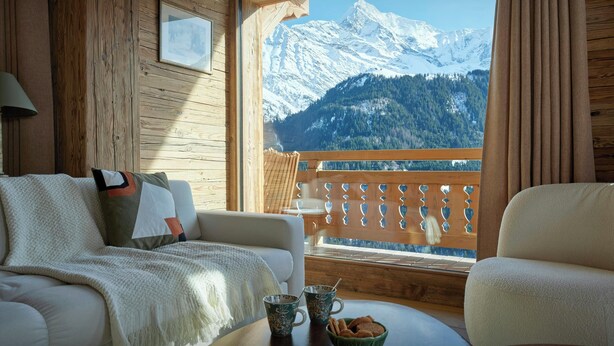 Ein gemütliches Zimmer mit Holzvertäfelung, einer Couch, einem Tisch mit Tasse und Keksen sowie einem großen Fenster mit Blick auf die Berge.