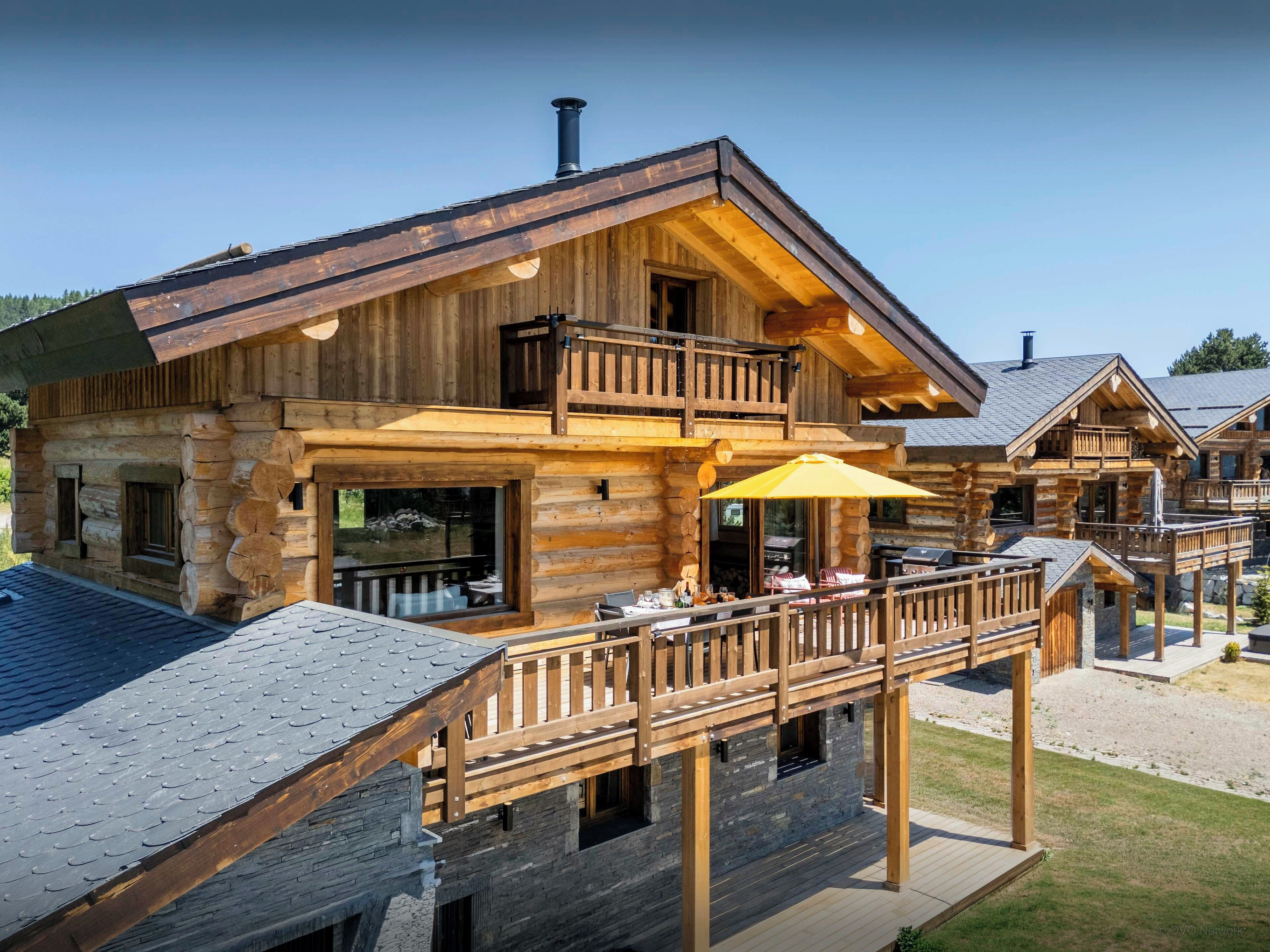 A log cabin with a balcony, outdoor seating, and a stone fireplace.