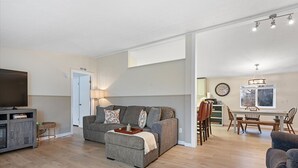 Uma sala de estar moderna com um sofá cinzento, um banquinho e uma televisão de tela plana. Há uma área de jantar com uma mesa e cadeiras de madeira, e uma cozinha visível ao fundo.
