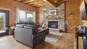 Una sala de estar acogedora con chimenea de piedra, vigas de madera, un sofá de cuero y un ventanal con vistas a la vegetación.