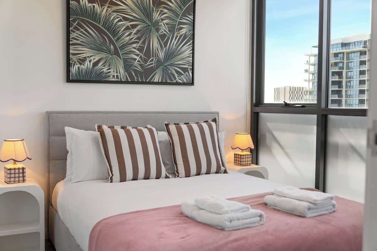 Een hotelkamer met een bed, kussens, een roze deken en uitzicht op gebouwen door grote ramen.