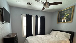 Une chambre à coucher avec un lit, un ventilateur de plafond, une fenêtre avec des rideaux, une commode et un tableau accroché au mur.