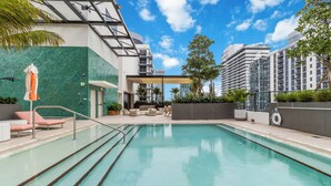 A swimming pool with lounge chairs and a covered seating area, surrounded by greenery and modern buildings. - Modern 1BR at The Crosby - Rooftop Pool, Gym & Walk to Bayside, Miami (Miami)