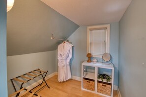 Ein Zimmer mit schrägem Dach, einem Wäscheständer, einem Bademantel an einem Gestell, einem kleinen weißen Beistelltisch mit einer Lampe und einem Fenster mit Jalousien.