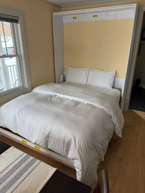 Un lit avec une literie blanche, dans une pièce dotée d’une fenêtre, d’une porte et d’un plancher en bois.