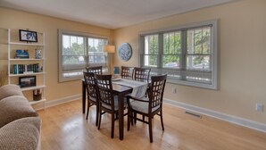 A dining area with a wooden table and chairs, a bookshelf, and large windows with blinds.