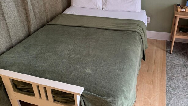 Een enkel bed met een groene deken, een houten nachtkastje met een lamp en een raam met jaloezieën.