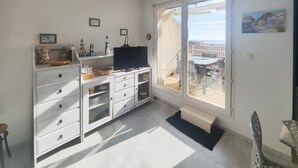 Una habitación con un amplio ventanal corredera que da a un balcón con mesa y sillas. Se observa un mueble blanco con cajones y estantes, un televisor encima y varios objetos decorativos en los estantes.