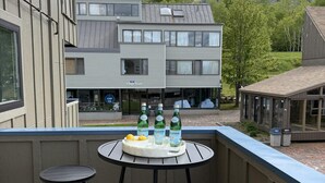 A balcony with two round tables and stools, a tray with bottles and lemons, and a view of a building with a sign 'Village South'.