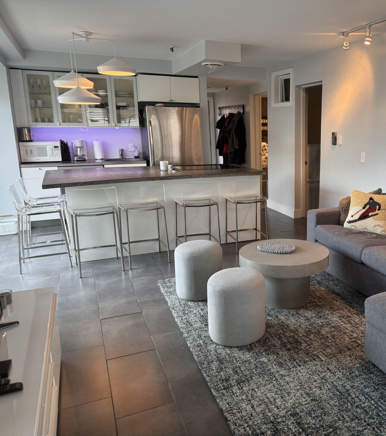 A modern kitchen with a central island, clear bar stools, and a grey sofa in a living area.