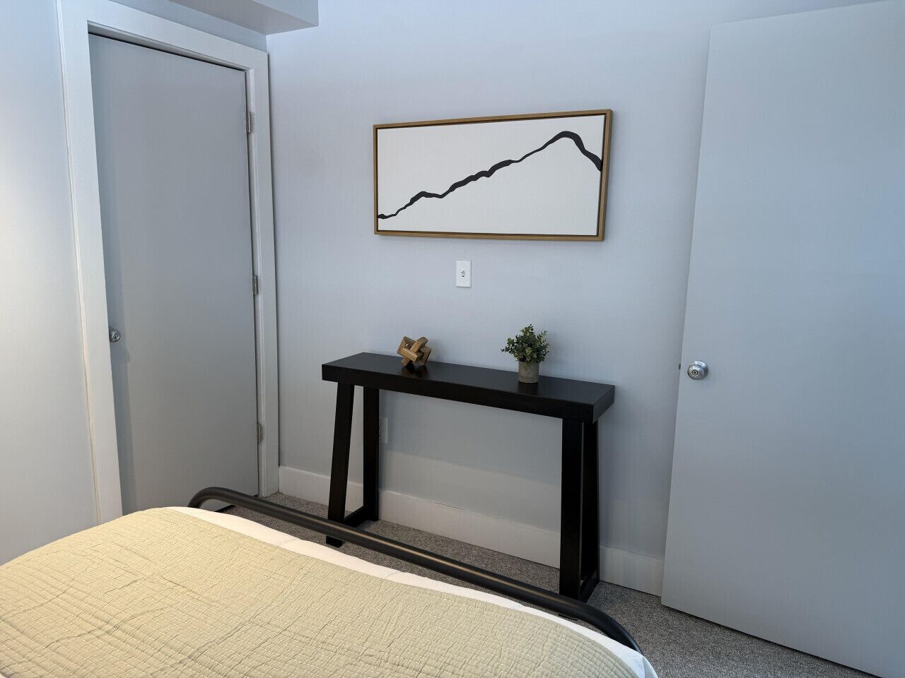 A bedroom with a bed, a black console table with a plant, and a framed line graph on the wall.