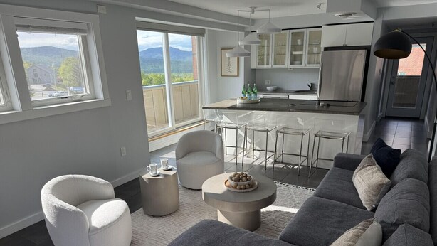 A modern living room with a grey sofa, a white armchair, and a round coffee table. There is a kitchen in the background with white cabinets and a stainless steel refrigerator.