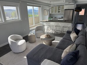A modern living room with a grey sofa, a white armchair, and a round coffee table. There is a kitchen in the background with white cabinets and a stainless steel refrigerator.