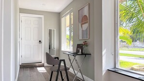 A hallway with a white door, a chair, a small table with a vase of flowers, and a window with a view of greenery. - Tranquil 4-bedroom house with heated pool and spa in Fort Lauderdale (Fort Lauderdale)