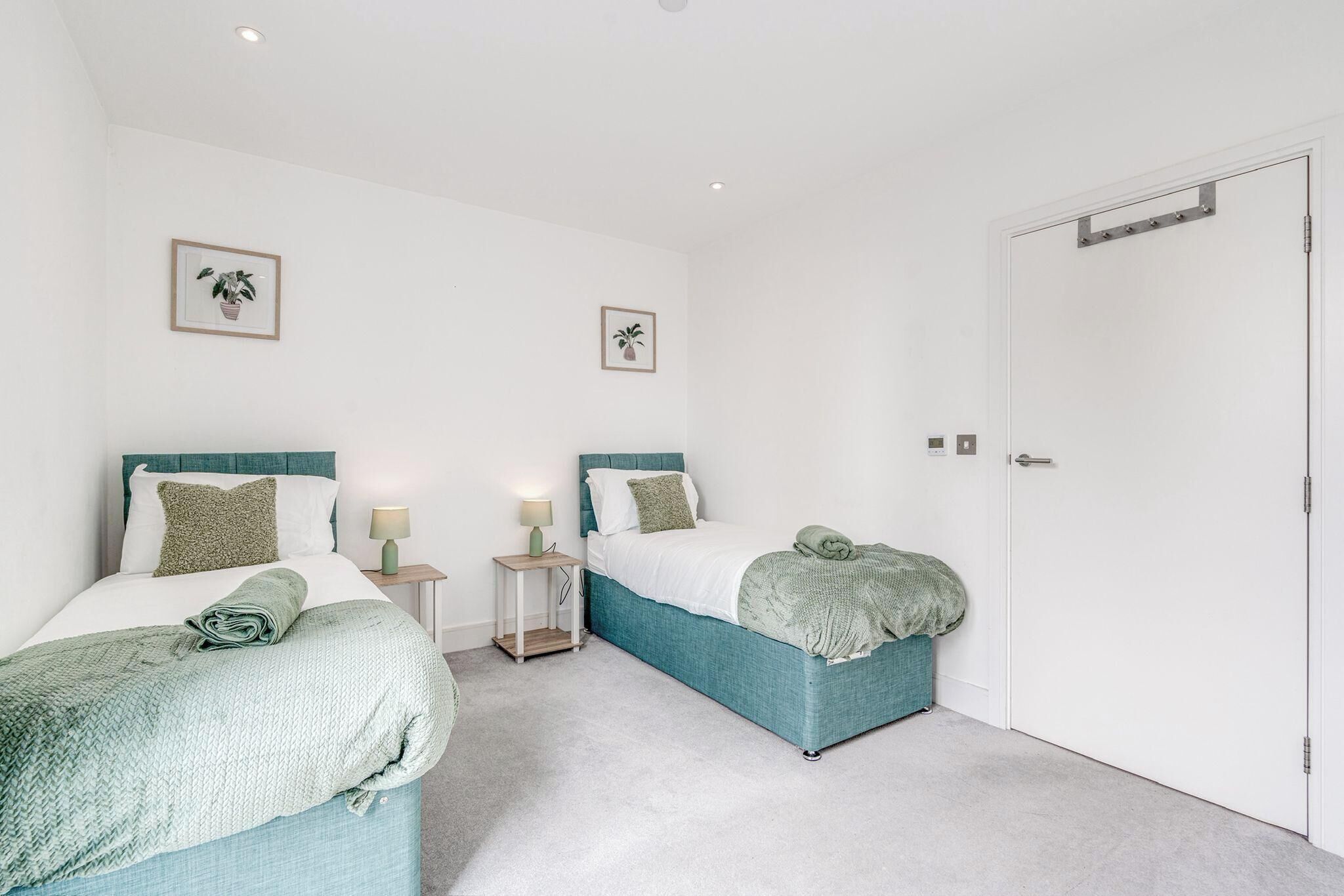 A bedroom with two beds, each with a teal headboard, white bedding, and green throw pillows. There are two bedside tables with lamps, and the room has white walls and a white door.