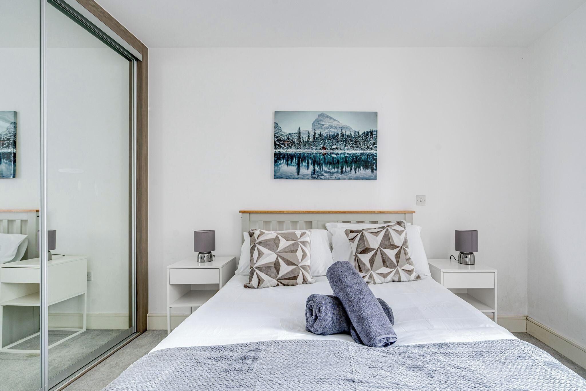 A modern hotel room with a large bed, two bedside tables, a glass partition, and a painting of a snowy landscape.