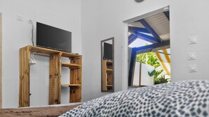 A bedroom with a bed, a wooden shelf with a TV, a mirror, and a window with a view of greenery. - TERAKOTA Village Créole jacuzzis-plage (Deshaies)