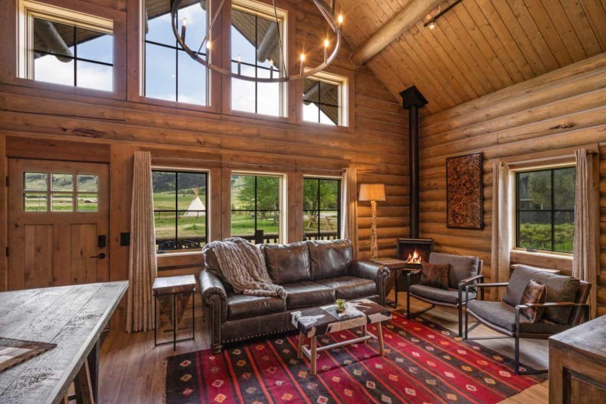A log cabin interior with a leather sofa, armchairs, a fireplace, and large windows overlooking a green landscape.