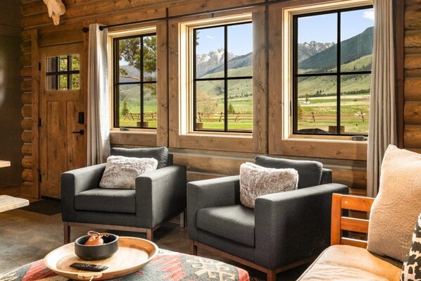 A log cabin room with two grey armchairs, a wooden coffee table, and a view of mountains through large windows.