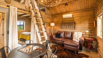 Interior de una cabaña de troncos con escalera de madera, sofá de cuero, mesa de comedor y una araña.