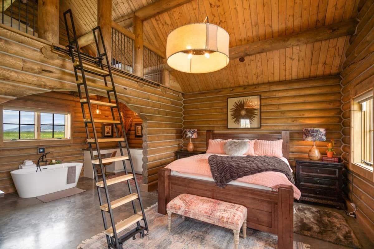 A log cabin bedroom with a wooden bed, a ladder, a bathtub, and a large window.