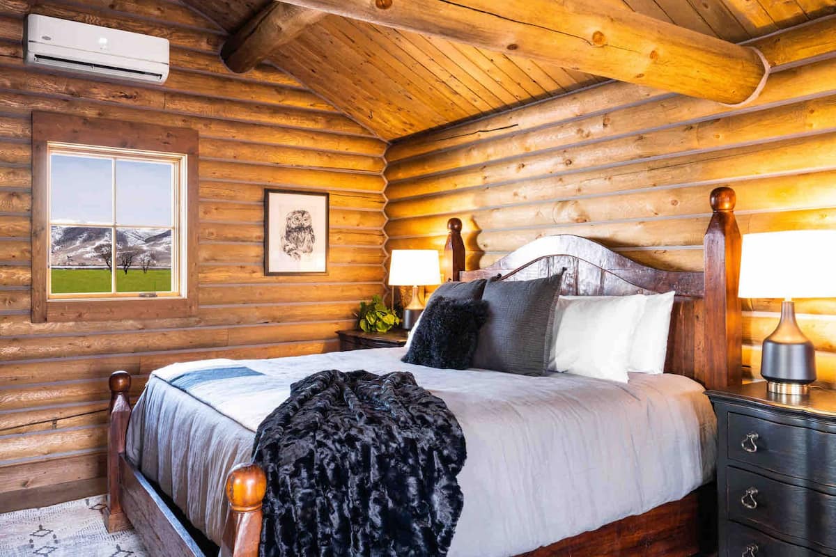 A log cabin bedroom with a bed, bedside tables, a window with a view, and an air conditioning unit.
