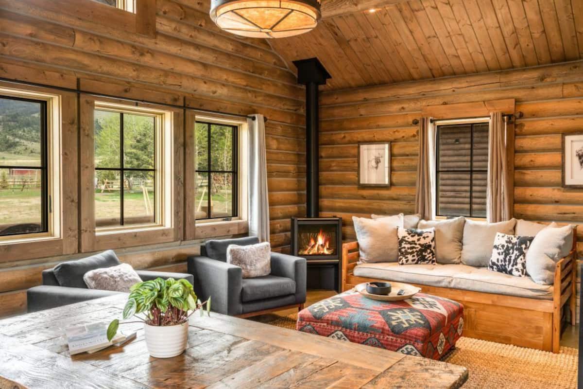A cozy log cabin interior with a fireplace, wooden walls, and large windows offering a view of the outdoors.