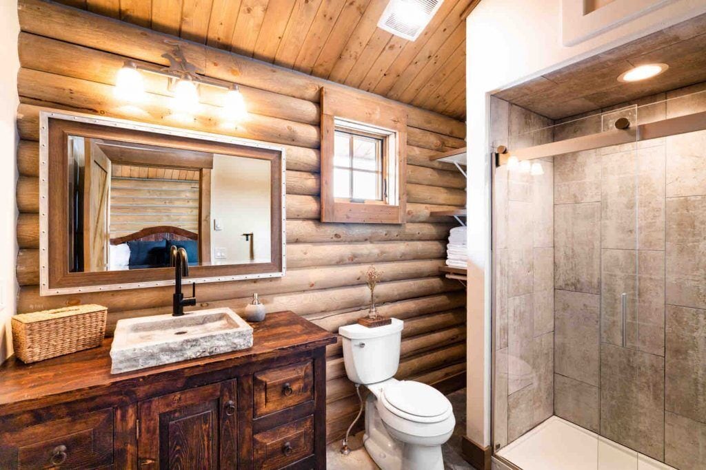 Een houten badkamer in een blokhutstijl, met een houten wastafel, een grote spiegel, een toilet en een douchehoek.