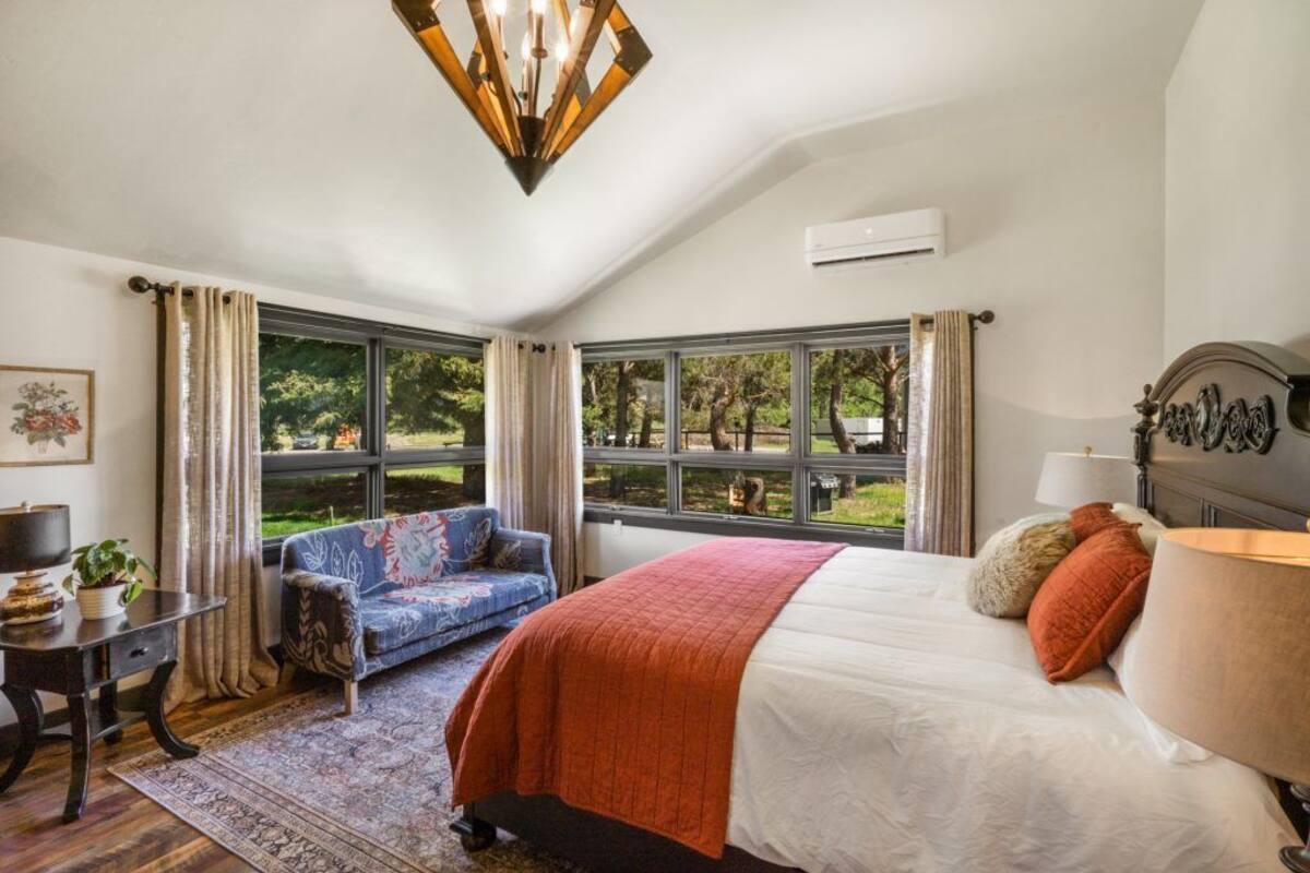A bedroom with a large bed, a sofa, and a desk. There are large windows with curtains, a chandelier, and an air conditioning unit.