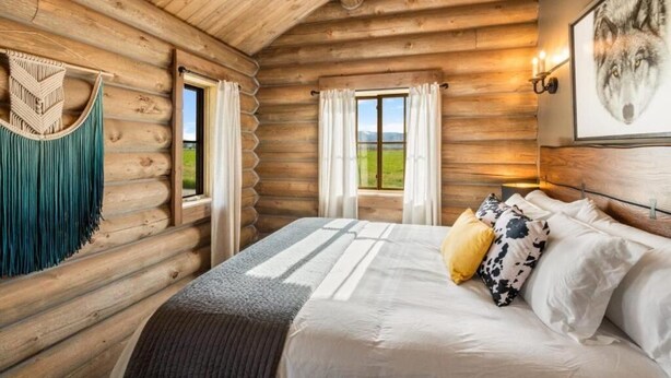 A log cabin bedroom with a bed, curtains, a wall-mounted light, and a framed picture. - Copper Rose Ranch (Livingston)