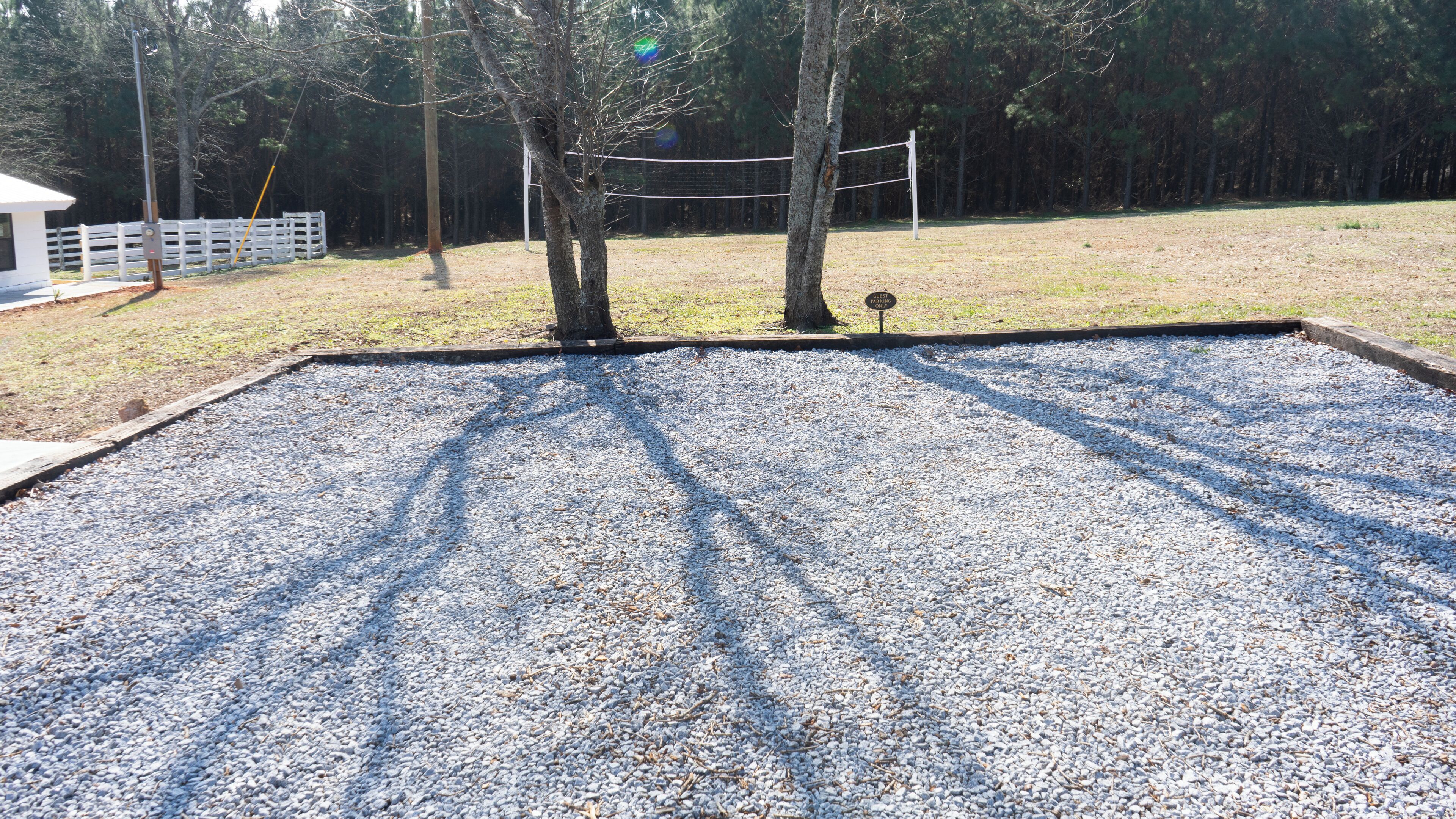 A gravel volleyball court with a white fence and trees in the background.