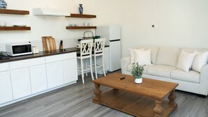 A compact kitchen with white cabinets, a microwave, and a toaster. There is a wooden coffee table with a plant, a white sofa, and a white chair.