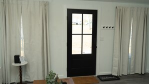 A room with a black door, white walls, and beige curtains. There is a small table with a plant and a black object on it.