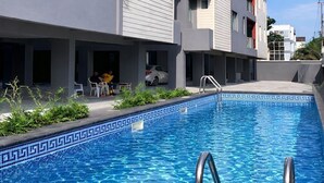 A modern building with a swimming pool, outdoor seating, and a clear blue sky. - Hibernate - 2 Bedroom Apartment - VI (Lagos)