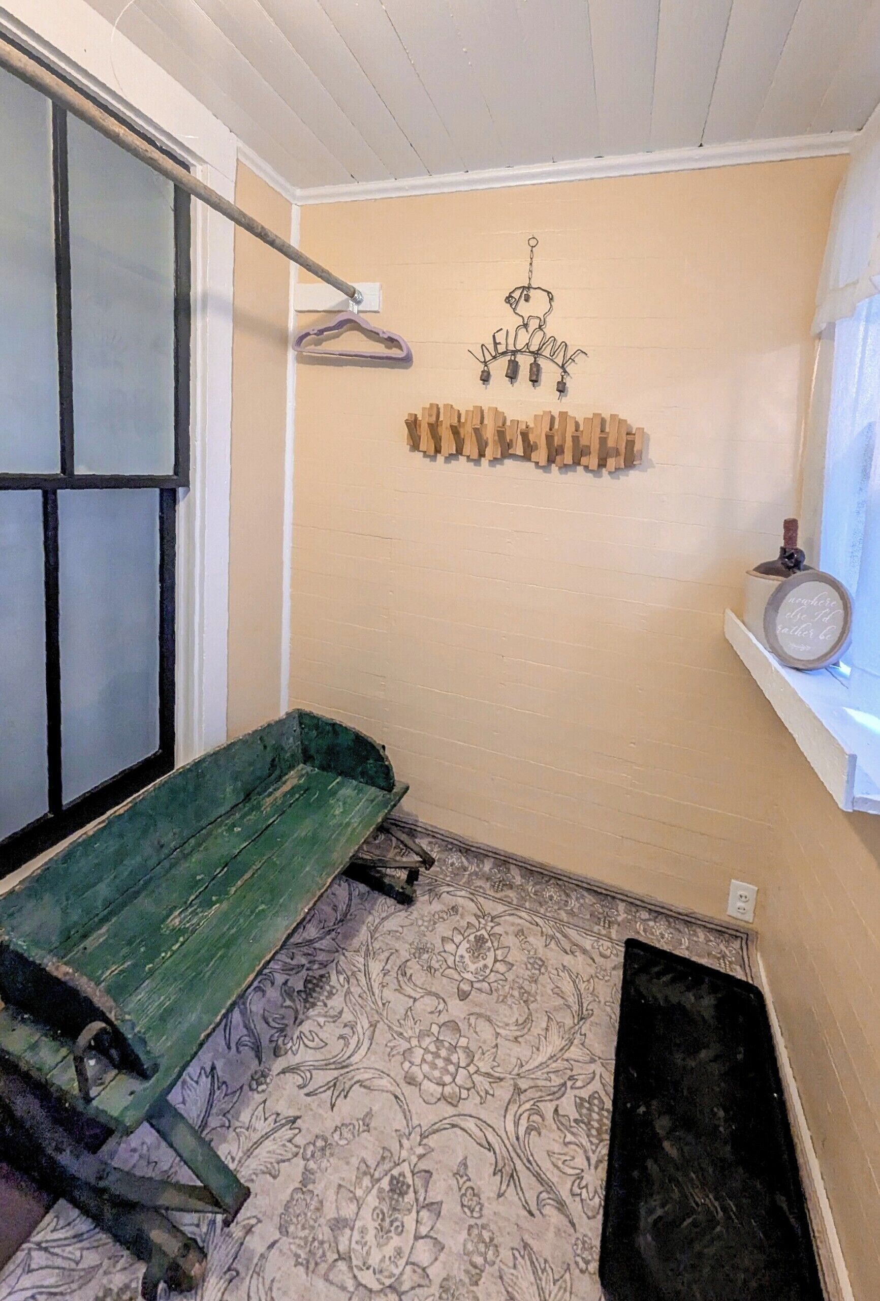 A room with a green bench, a hanging light fixture, a wall-mounted coat rack, and a small shelf with a clock.